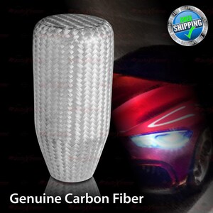 May include: A white carbon fiber shift knob with a woven pattern. The text "Genuine Carbon Fiber" is displayed at the bottom of the image. The background features a red car with bright headlights.