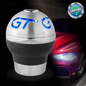 May include: A silver and black car gear shift knob with the letters "GT" illuminated in blue. The knob has a leather-like texture on the lower portion and a shiny metal finish on the top and bottom.
