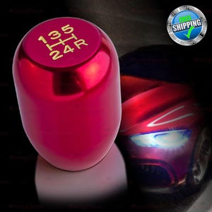 May include: A shiny red shift knob with gold lettering indicating gear positions. The knob has a smooth, rounded shape and is designed for a vehicle's manual transmission. The background features a blurred image of a red car.