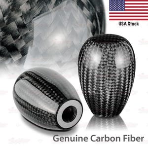 May include: Two carbon fiber shift knobs. One is oval-shaped, the other is round. Both have a glossy finish and a woven pattern. The text "Genuine Carbon Fiber" is at the bottom. The American flag and "USA Stock" are in the upper right corner.