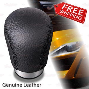 May include: A black, leather shift knob with a stitched seam and a silver base. The text "Genuine Leather" is at the bottom of the image.