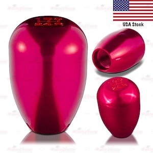 May include: A set of three shiny, red shift knobs. The main knob is oval-shaped with the gear pattern 1 3 5 R 2 4 on top. The other two knobs are angled and show the threaded interior. The USA Stock flag is in the background.