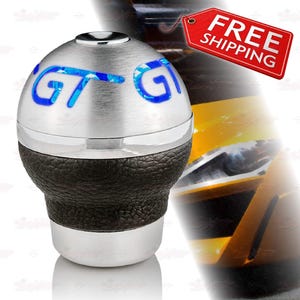 May include: A silver and black car gear shift knob with the letters "GT" in blue. The knob has a textured black leather base and a shiny silver top. The text "FREE SHIPPING" is on a red tag.