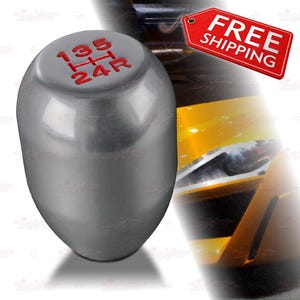 May include: Silver-colored car shift knob with red gear numbers. The top of the knob displays the gear pattern: 1, 3, 5, H, 2, 4, and R. The knob has a smooth, rounded shape and is designed for a vehicle's manual transmission.