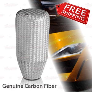 May include: A silver and gray shift knob with a woven carbon fiber pattern. The shift knob has a rounded top and a tapered bottom. The text "Genuine Carbon Fiber" is at the bottom of the image.