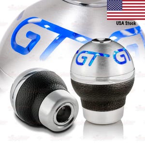 May include: Two silver and black car shift knobs with the letters "GT" illuminated in blue. The knobs have a textured black leather grip and a silver base. The image also includes a USA flag.