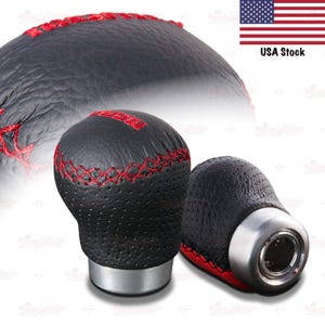 May include: Black leather shift knobs with red stitching. The knobs have a textured surface and a silver base. The image also includes a blurred shift knob in the background and a USA flag.