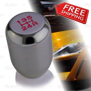 May include: A silver-colored shift knob with red gear markings. The top of the knob displays the gear pattern: 1, 3, 5, and 2, 4, R. The knob has a smooth, rounded shape and is designed for a vehicle's manual transmission.