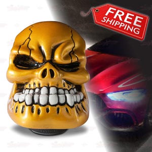 May include: A gold-colored skull-shaped car accessory with black eye sockets and white teeth. The skull has cracks across the top and is set against a blurred background with a red object. A red tag with the words "FREE SHIPPING" is visible.