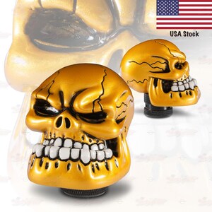 May include: Gold skull-shaped car shift knobs. The knobs have a metallic gold finish with black detailing in the eye sockets and cracks. The teeth are white. The image includes the text "USA Stock".
