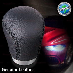 May include: Black leather gear shift knob with visible stitching. The knob has a silver base and the words "Genuine Leather" are printed at the bottom. A red car is blurred in the background.
