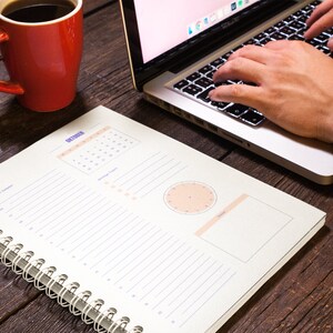 Pu&ograve; includere: Un'agenda bianca a spirale con un calendario per ottobre, un orologio e una sezione di note vuota. L'agenda &egrave; su un tavolo di legno con un laptop e una tazza di caff&egrave; rossa.