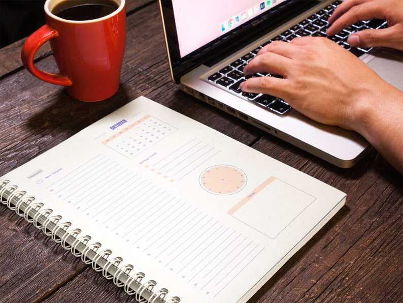 Pu&ograve; includere: Un'agenda bianca a spirale con un calendario per ottobre, un orologio e una sezione di note vuota. L'agenda &egrave; su un tavolo di legno con un laptop e una tazza di caff&egrave; rossa.