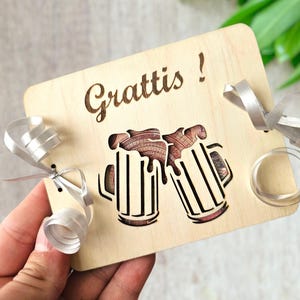 May include: Wooden plaque with the word "Grattis!" and a laser-cut design of two beer mugs clinking. The plaque is tied with a silver ribbon. The mugs are filled with a brown liquid.