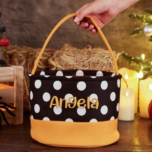 May include: A black and white polka dot trick-or-treat bucket with a yellow handle and a yellow bottom. The bucket is personalized with the name "Angela" in yellow embroidery.