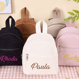 May include: Five mini corduroy backpacks in different colors, with personalized names embroidered on the front. The backpacks are black, brown, white, gray, and pink. The names are Rhoda, Paula, Becky.