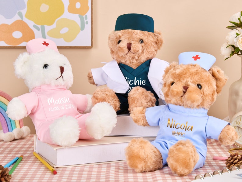 May include: Three plush teddy bears dressed as nurses. The bear on the left is wearing a pink uniform with the name "Maisie" and "Class of 2024" embroidered on it. The bear in the middle is wearing a blue uniform with a white doctor's coat and the name "Richie" embroidered on it. The bear on the right is wearing a blue uniform with the name "Nicola" embroidered on it.