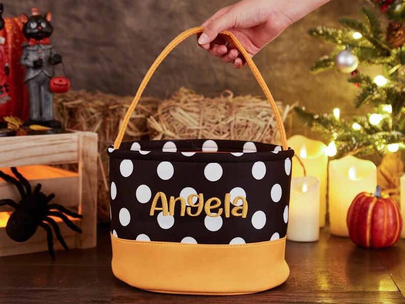 May include: A black and white polka dot trick-or-treat bucket with a yellow handle and a yellow bottom. The bucket is personalized with the name "Angela" in yellow embroidery.