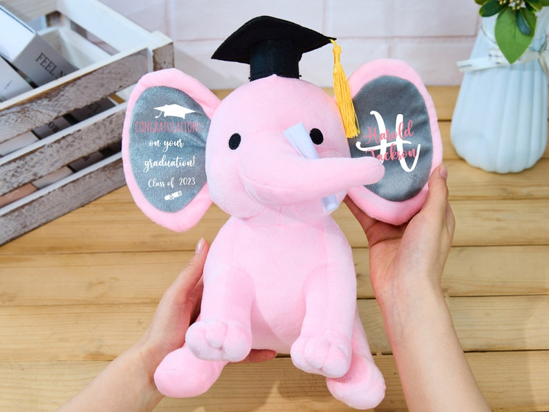 Puede incluir: Elefante de peluche rosa con un gorro de graduaci&oacute;n y sosteniendo un diploma. Las orejas del elefante est&aacute;n personalizadas con el texto "&iexcl;Felicidades por tu graduaci&oacute;n! Clase de 2023" y "Harold Jackson".