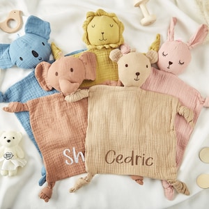Könnte beinhalten: Fünf bunte Baby-Sicherheitsdecken mit Tiergesichtern und bestickten Namen. Die Decken sind aus weichem, strukturiertem Stoff und haben geknüpfte Ecken. Die Tiere sind ein Koala, ein Elefant, ein Löwe, ein Bär und ein Kaninchen.