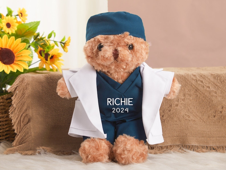 May include: A brown teddy bear wearing a white doctor's coat and a blue scrub hat and scrubs. The scrubs have the name "RICHIE" and the year "2024" embroidered on the front.