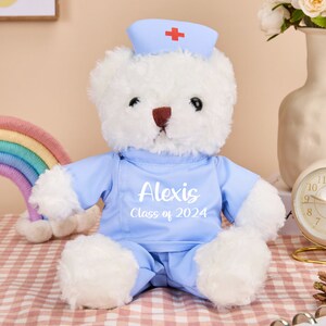 May include: A white teddy bear wearing a blue nurse's uniform with a red cross on the hat. The bear has a name tag that says "Alexis Class of 2024".