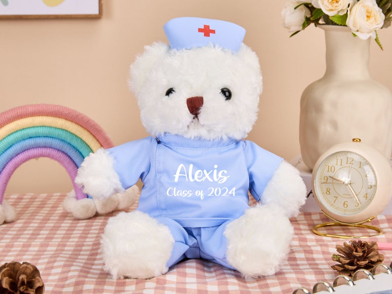 May include: A white teddy bear wearing a blue nurse's uniform with a red cross on the hat. The bear has a name tag that says "Alexis Class of 2024".