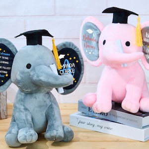 Puede incluir: Dos peluches de elefante gris y rosa con gorros de graduaci&oacute;n y diplomas. El elefante gris tiene un parche negro en la oreja con el texto "CHASE YOUR dreams NEVER STOP TRYING NEVER STOP LEARNING LIVE life TO THE FULLEST AND GIVE IT NOTHING BUT YOUR best" y el texto "MANDA CLARKE". El elefante rosa tiene un parche gris en la oreja con el texto "Belinda Davies" y el texto "HAPPY GRADUATION! Class of 2023".