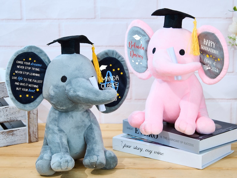 Puede incluir: Dos peluches de elefante gris y rosa con gorros de graduaci&oacute;n y diplomas. El elefante gris tiene un parche negro en la oreja con el texto "CHASE YOUR dreams NEVER STOP TRYING NEVER STOP LEARNING LIVE life TO THE FULLEST AND GIVE IT NOTHING BUT YOUR best" y el texto "MANDA CLARKE". El elefante rosa tiene un parche gris en la oreja con el texto "Belinda Davies" y el texto "HAPPY GRADUATION! Class of 2023".