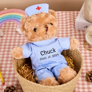 May include: A light blue teddy bear wearing a nurse's hat and scrubs. The bear has a name tag that says "Chuck Class of 2024".