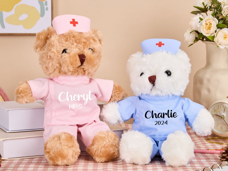 May include: Two plush teddy bears dressed as nurses. The brown bear is wearing a pink uniform with the name "Cheryl HNS" embroidered on the front. The white bear is wearing a blue uniform with the name "Charlie 2024" embroidered on the front.