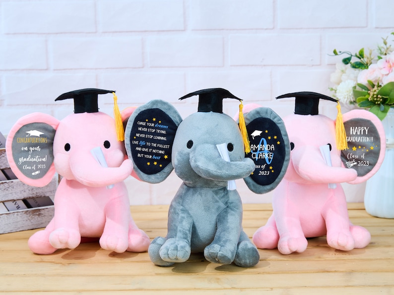 Puede incluir: Tres peluches de elefante con gorros de graduaci&oacute;n y sosteniendo carteles con mensajes personalizados. Los elefantes son rosas, grises y rosas. Los carteles dicen "&iexcl;Felicidades por tu graduaci&oacute;n! Clase de 2023", "Persigue tus sue&ntilde;os, nunca dejes de intentar, nunca dejes de aprender, vive la vida al m&aacute;ximo y no le des nada menos que tu mejor esfuerzo", y "Amanda Clarke Clase de 2023".