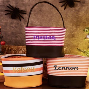 May include: Three striped Halloween trick-or-treat bags with black handles. The bags are personalized with the names "Marian", "Kelsey", and "Lennon".