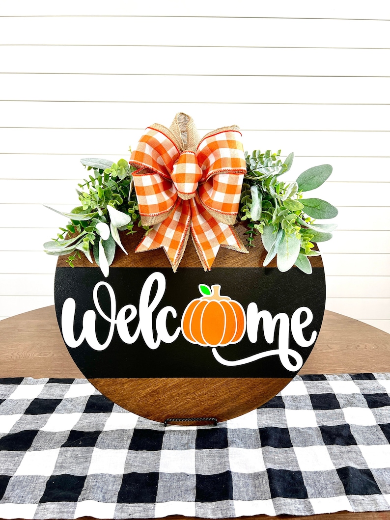 May include: Round wooden welcome sign with a black and brown background. The word "Welcome" is in white script, with an orange pumpkin graphic. The sign is decorated with greenery and a large orange and white plaid bow.