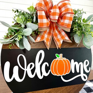 May include: A round wooden welcome sign with a black stripe and the word "Welcome" in white script. An orange pumpkin graphic is next to the word "Welcome". The sign is decorated with a large orange and white checkered bow and greenery.