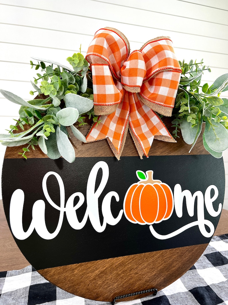 May include: A round wooden welcome sign with a black stripe and the word "Welcome" in white script. An orange pumpkin graphic is next to the word "Welcome". The sign is decorated with a large orange and white checkered bow and greenery.