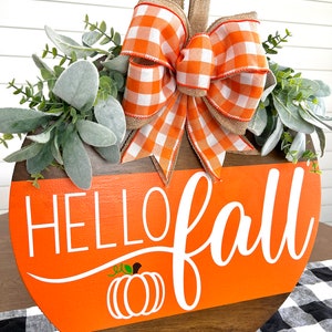 May include: A round wooden sign with the words "HELLO fall" in white script, an orange background, and a pumpkin graphic. The sign is decorated with a plaid orange and white bow, greenery, and a burlap hanger.