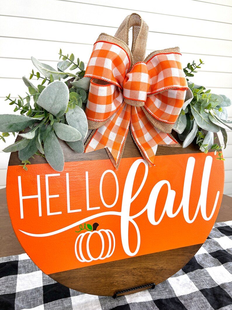 May include: A round wooden sign with the words "HELLO fall" in white script, an orange background, and a pumpkin graphic. The sign is decorated with a plaid orange and white bow, greenery, and a burlap hanger.