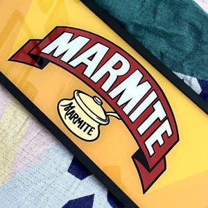 Puede incluir: Una impresión enmarcada del logotipo de la marca Marmite. El logotipo presenta una pancarta roja y blanca con la palabra "Marmite" en letras blancas. Una olla amarilla con la palabra "Marmite" en letras negras está en el centro de la pancarta. El fondo es amarillo.