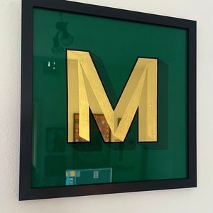 May include: A gold letter M in a black frame against a green background.