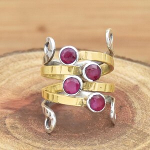 Può includere: Un anello multistrato caratterizzato da una fascia dorata a spirale impreziosita da quattro rubini rotondi, sfaccettati e di colore rosa intenso. La struttura presenta elementi metallici argentati alle estremità superiore e inferiore.
