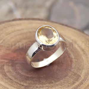 May include: A silver ring featuring a large, oval-cut, yellow gemstone. The gemstone is set in a polished silver band, creating a classic and elegant design. The ring is displayed on a wooden surface.
