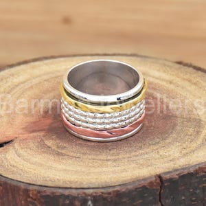 Può includere: Un anello girevole in argento, oro e rame con una fascia testurizzata. L'anello è composto da più fasce che girano liberamente.