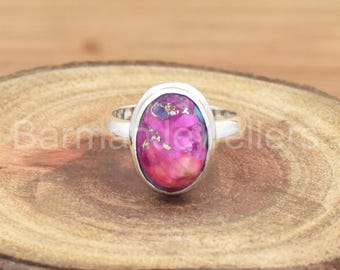 Puede incluir: Un anillo de plata con una piedra ovalada rosa, azul y dorada. El anillo está colocado sobre una superficie de madera.