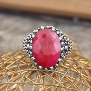 Può includere: Un anello d'argento con una grande gemma di rubino ovale. Il rubino è di un rosso brillante ed è incastonato in una fascia d'argento strutturata. L'anello è esposto su una superficie dorata e intrecciata.
