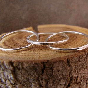 Könnte beinhalten: Drei silberne Ringe mit gehämmerten Texturen sind ineinander verschlungen und liegen auf einer Holzoberfläche. Die Ringe sind dünn und haben ein schlichtes, elegantes Design. Der Hintergrund ist verschwommen und neutral, wodurch der Schmuck hervorgehoben wird.
