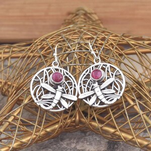 May include: Silver-toned dragonfly earrings with a round, openwork design. Each earring features a dragonfly and a round, red gemstone. The earrings are displayed on a gold-colored, star-shaped decorative object.