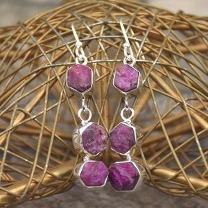 May include: Dangle earrings with hexagonal, deep pink gemstones set in silver-coloured metal. The earrings hang from a wire hook. The background is a gold-coloured, woven structure. The gemstones are approximately 1 cm in diameter.