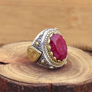 以下が含まれることがあります： 大きな楕円形の深い赤色の宝石が付いたシルバーリング。石とバンドの周りには、金と銀の複雑な装飾が施されています。リングは木製の表面に置かれています。