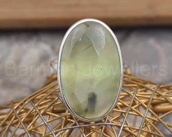 May include: A silver ring featuring a large, oval-cut, pale green gemstone. The gemstone has a translucent appearance with subtle internal inclusions. The ring is set on a gold-colored, woven surface.